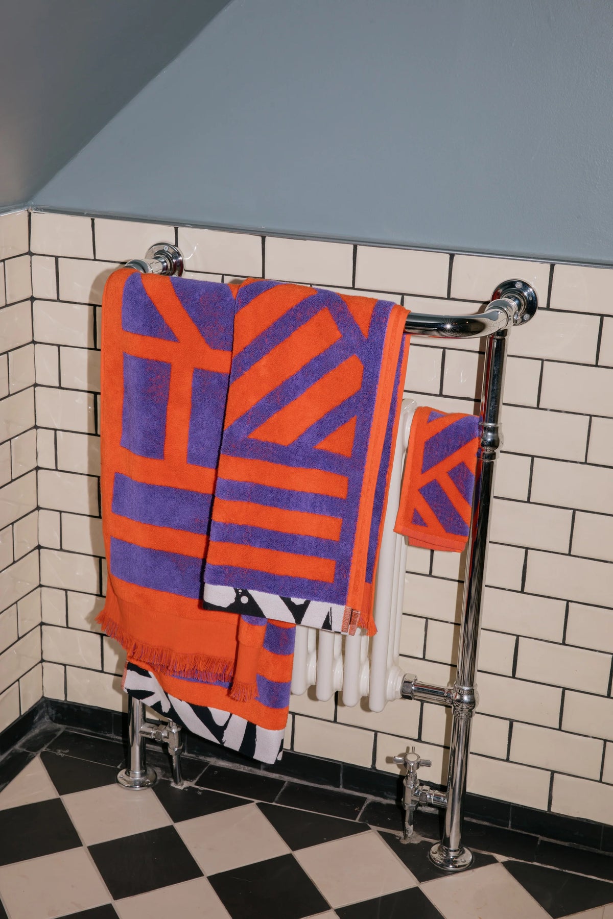 A set of orange and purple geometric patterned towels hanging on a chrome towel rail in a bathroom with white subway tiles and a black-and-white checkered floor.