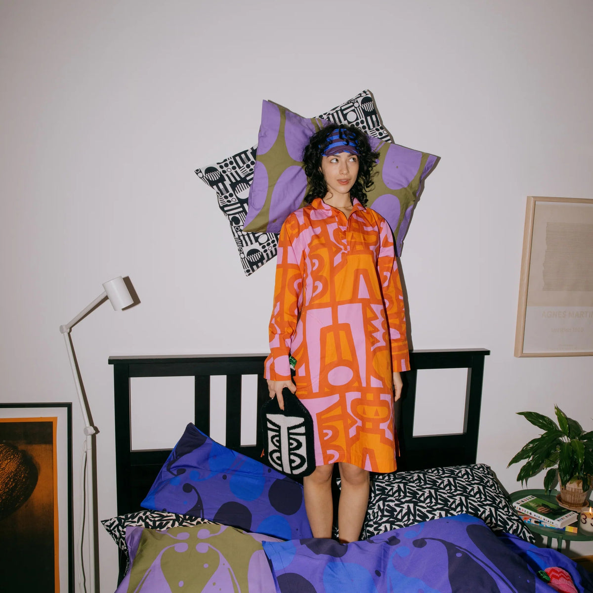 A person stands on a bed in a bedroom wearing a bright orange and pink abstract-patterned long-sleeve top and holding a black tote bag with white text. The bed is covered in matching multicolored patterned bedding, and geometric pillows in purple, black, and white are arranged behind them against a plain white wall.
