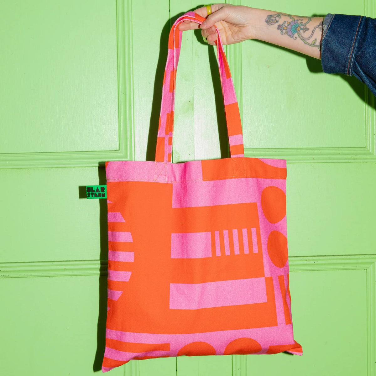 A hand with a tattoo holds up a bright orange and pink geometric patterned tote bag against a lime green paneled door.