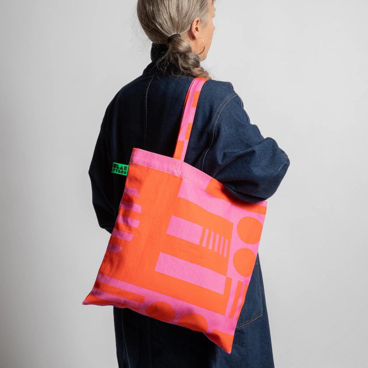 A woman viewed from behind wearing a dark denim jacket and carrying a bright pink and orange patterned tote bag over her shoulder, against a plain light gray studio background.