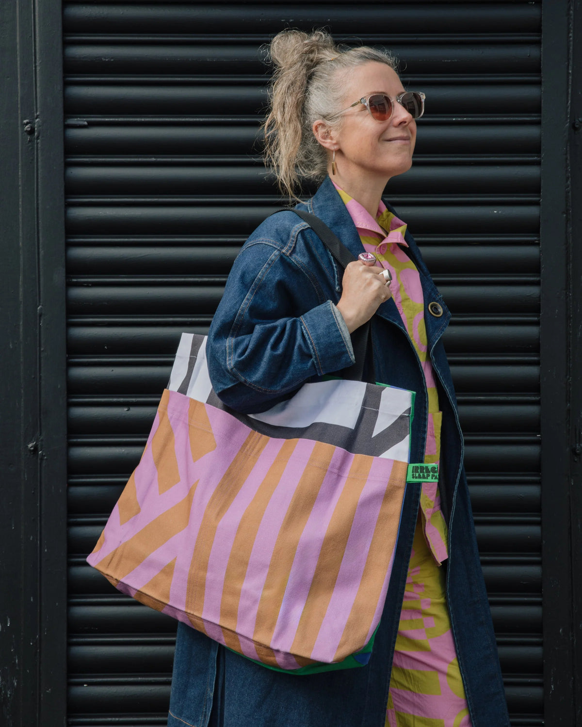 A woman wearing sunglasses and a denim jacket carries a large pink and orange striped tote bag with white and gray geometric accents, standing in front of a black metal shutter.
