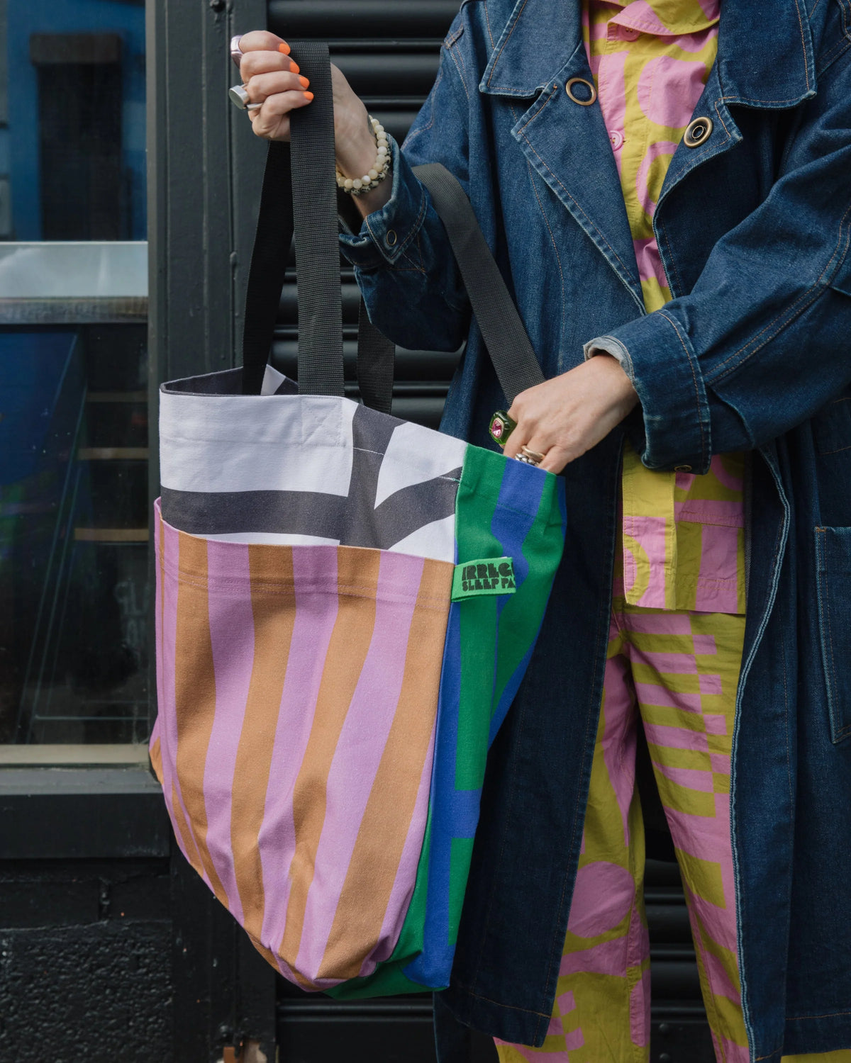 A person wearing a dark denim coat and a colorful pink, yellow, and green patterned outfit holds a large striped tote bag with black handles. The bag features vertical stripes in pink, beige, and orange on the front pocket, with white, black, green, and blue panels stacked behind it.