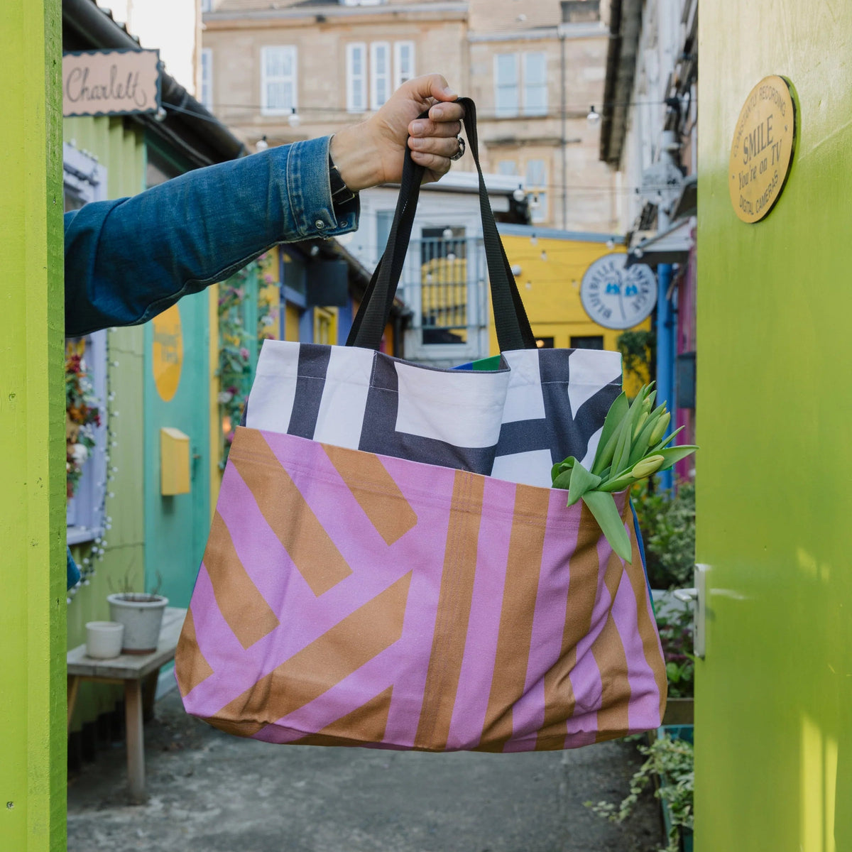 A person holds up a large tote bag with a bold geometric pattern in pink, brown, black, and white, filled with groceries in a colorful outdoor alleyway.