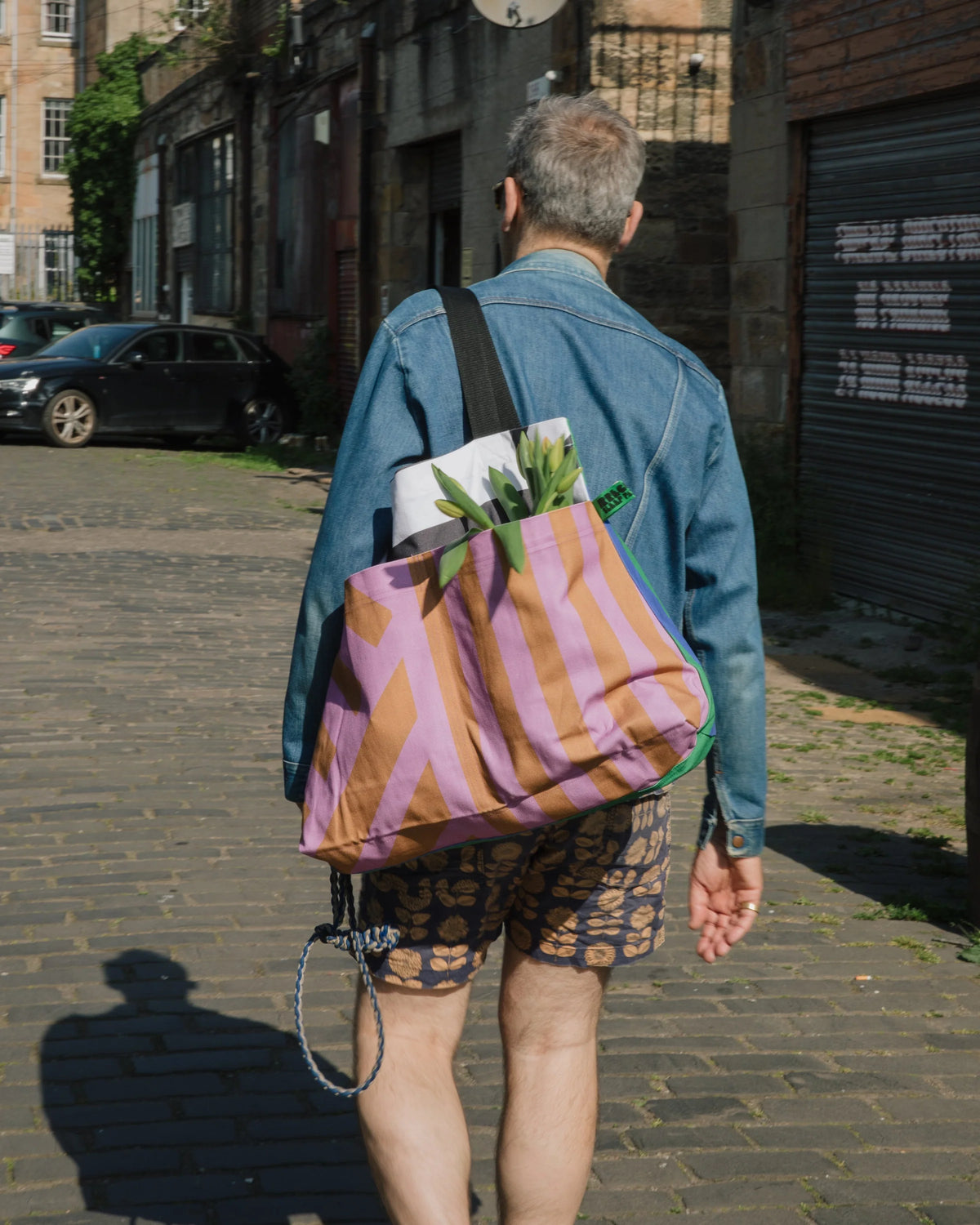 A person viewed from behind walking on a cobblestone street, wearing a blue denim jacket and patterned shorts, carrying a large pink and orange striped tote bag over their shoulder.