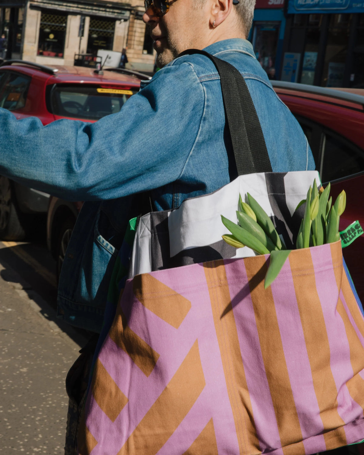 A man in a blue denim jacket and sunglasses carries a large striped reusable tote bag with pink, orange, and white diagonal patterns, filled with tulips, on a city street.