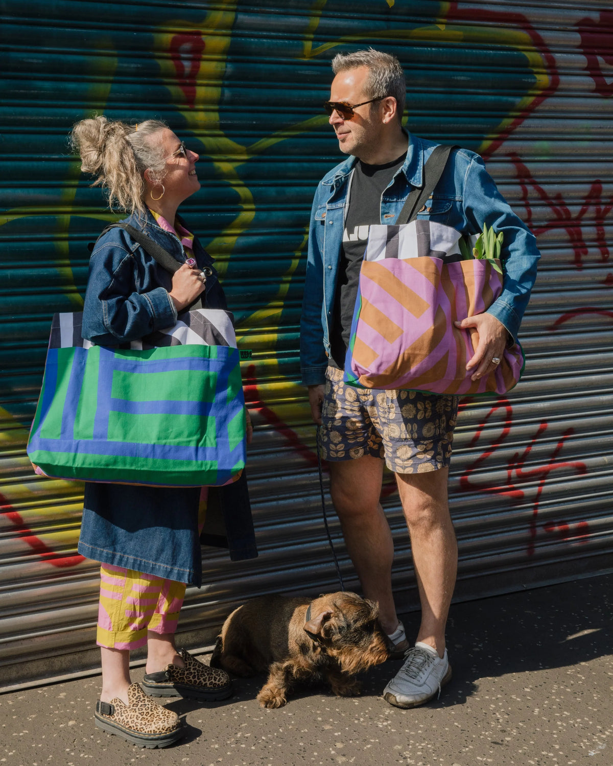 A man and a woman standing outdoors in front of a graffiti-covered metal shutter, each carrying a large striped tote bag. A small dog lies on the ground between them.