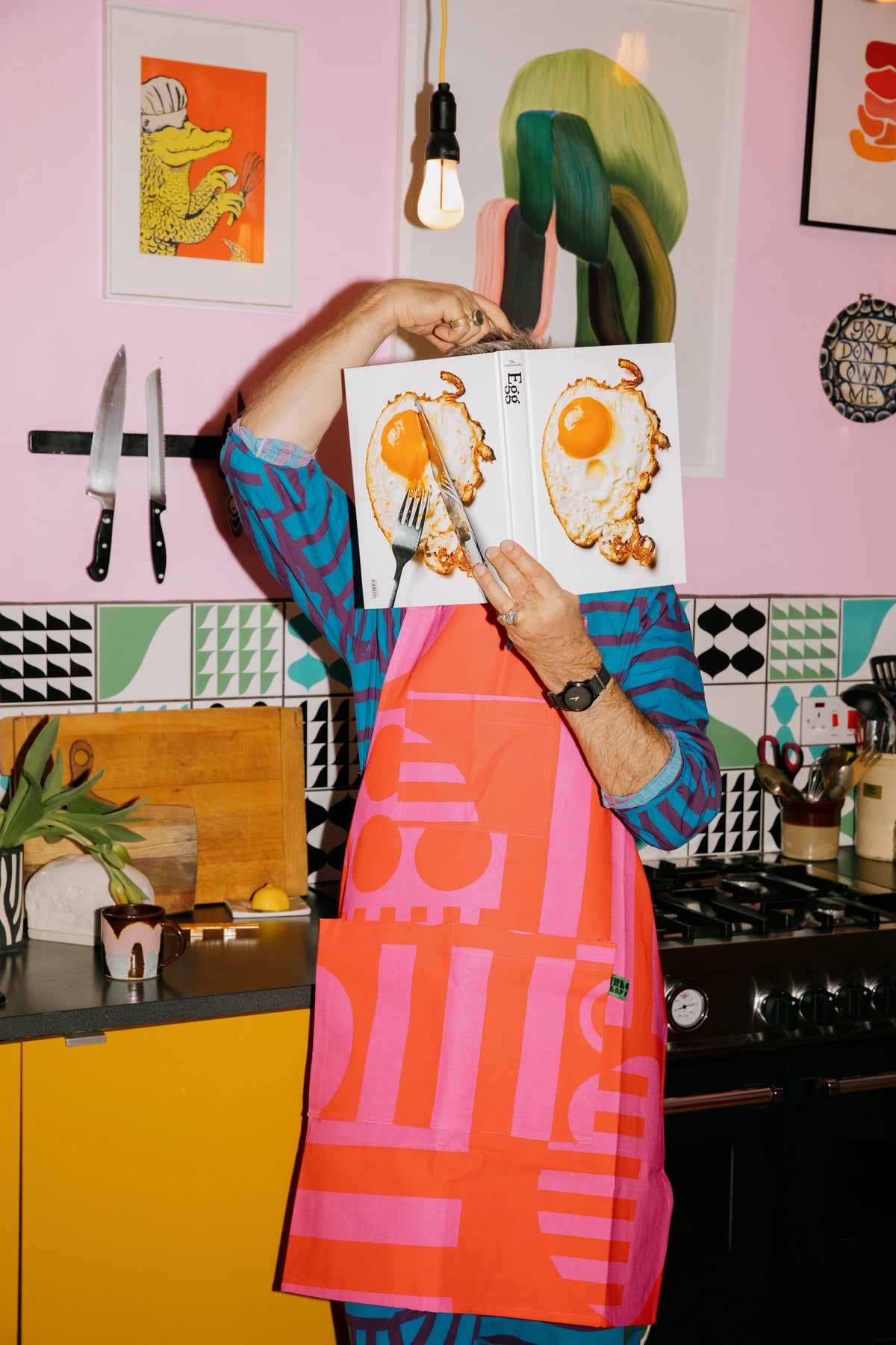 A person in a colorful kitchen setting holds up a cookbook with two illustrated eggs on the cover, wearing a bright pink and orange apron with a bold graphic pattern.