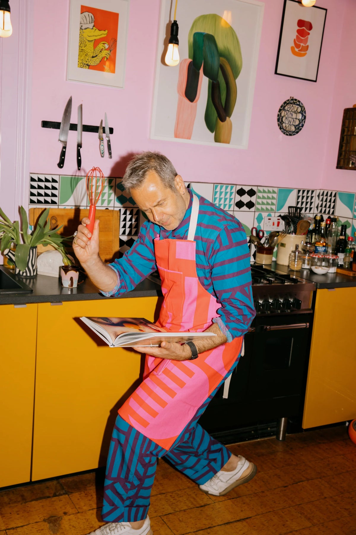 A man in a colorful kitchen setting, wearing a matching blue and pink patterned shirt and apron while holding a book and a red object.