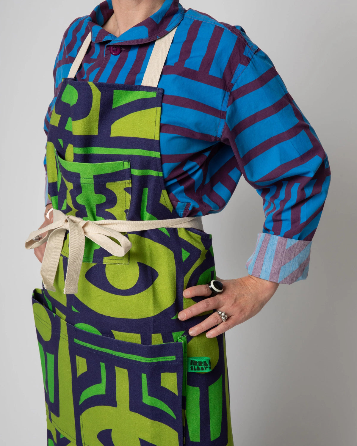 A person wearing a colorful apron over a blue and purple striped shirt, standing against a plain light gray background. The apron has a bold abstract pattern in lime green and dark purple with cream straps and waist tie.