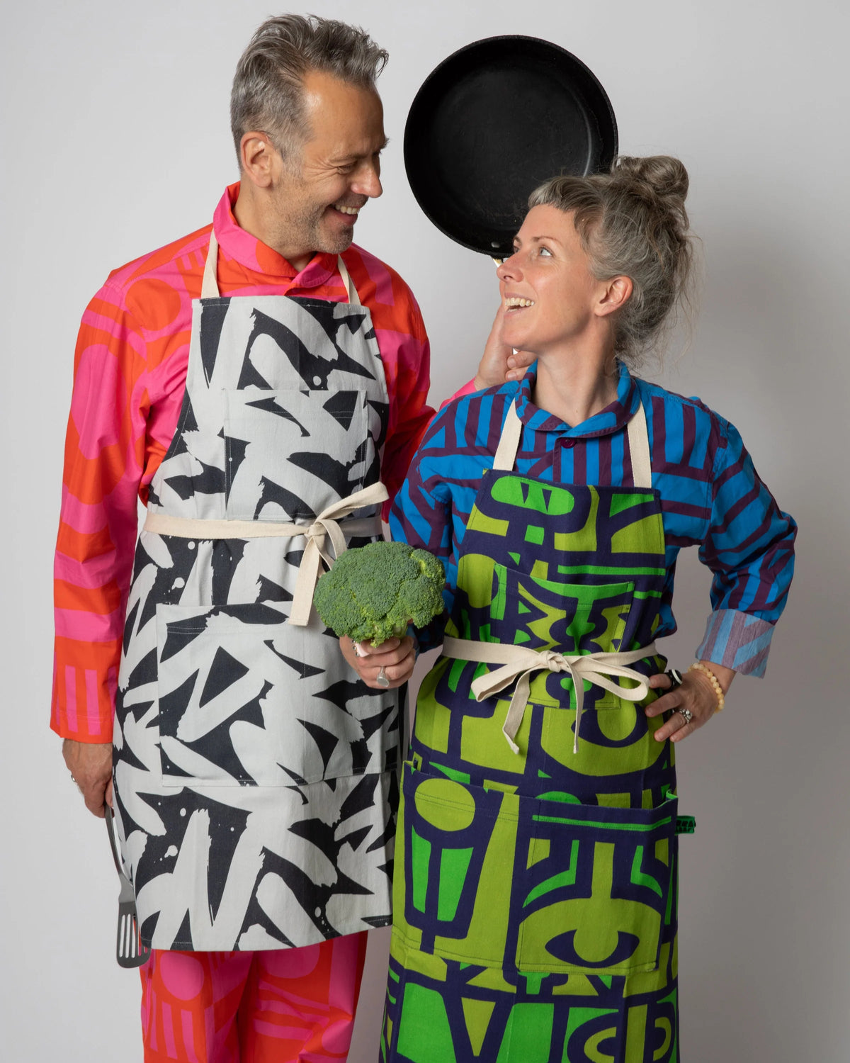 A man and a woman wearing colorful patterned aprons and shirts pose together against a light gray studio background. The man wears a black, white, and gray abstract patterned apron over a bright pink and orange striped shirt, while the woman wears a blue and green abstract patterned apron over a blue patterned shirt.