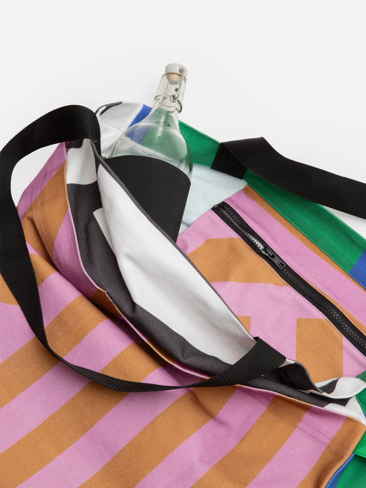 A close-up of a colorful striped tote bag with black straps, shown partially open with a clear glass bottle inside. The bag features bold diagonal stripes in pink, orange, white, black, green, and blue.