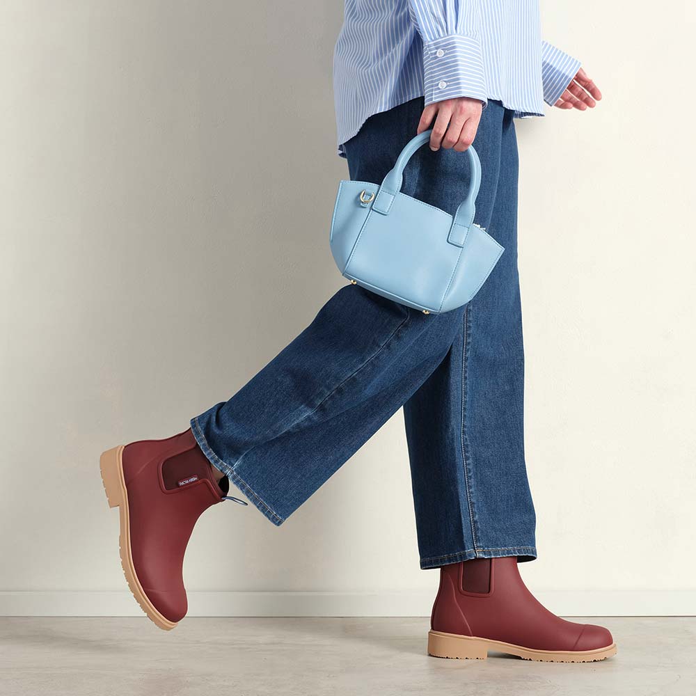 Person wearing maroon boots and blue jeans, holding a light blue handbag on a neutral background