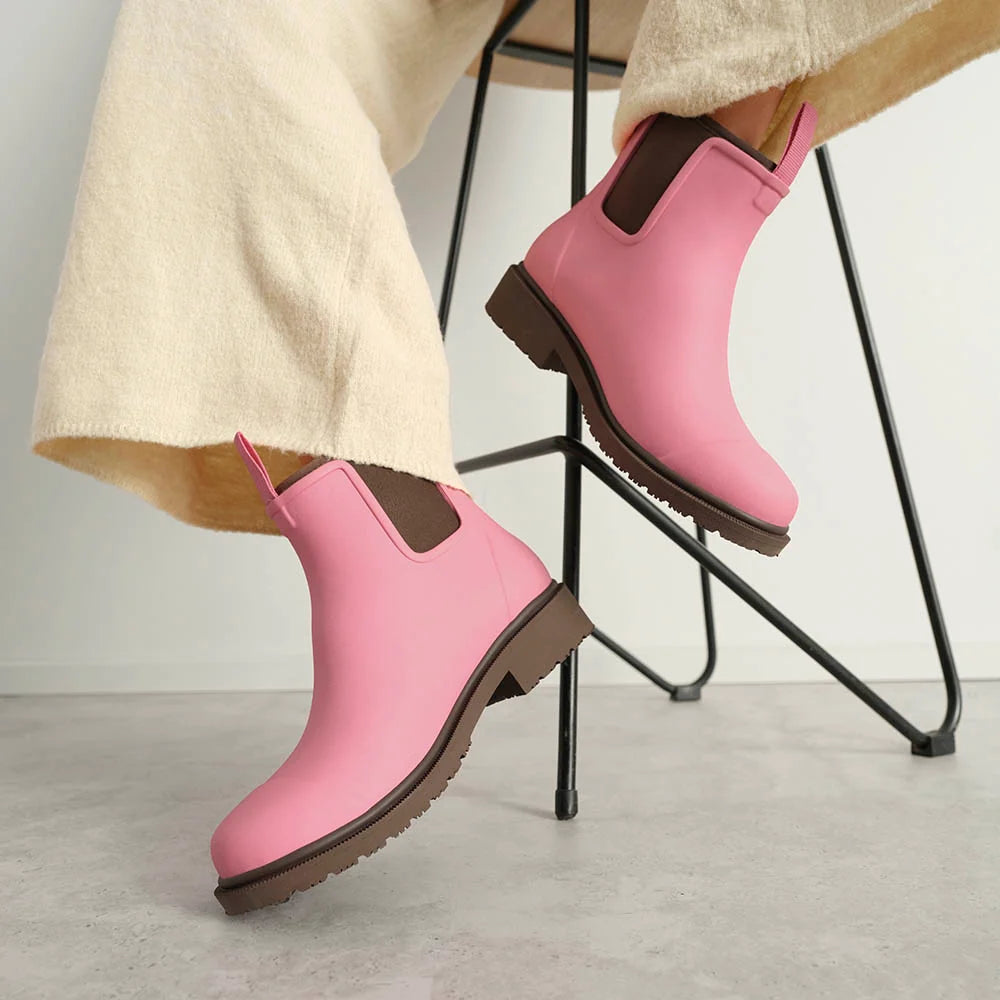 Pink Chelsea-style rain boots with brown trim and soles, shown on a person's feet with cream-colored trousers, seated on a black metal chair.