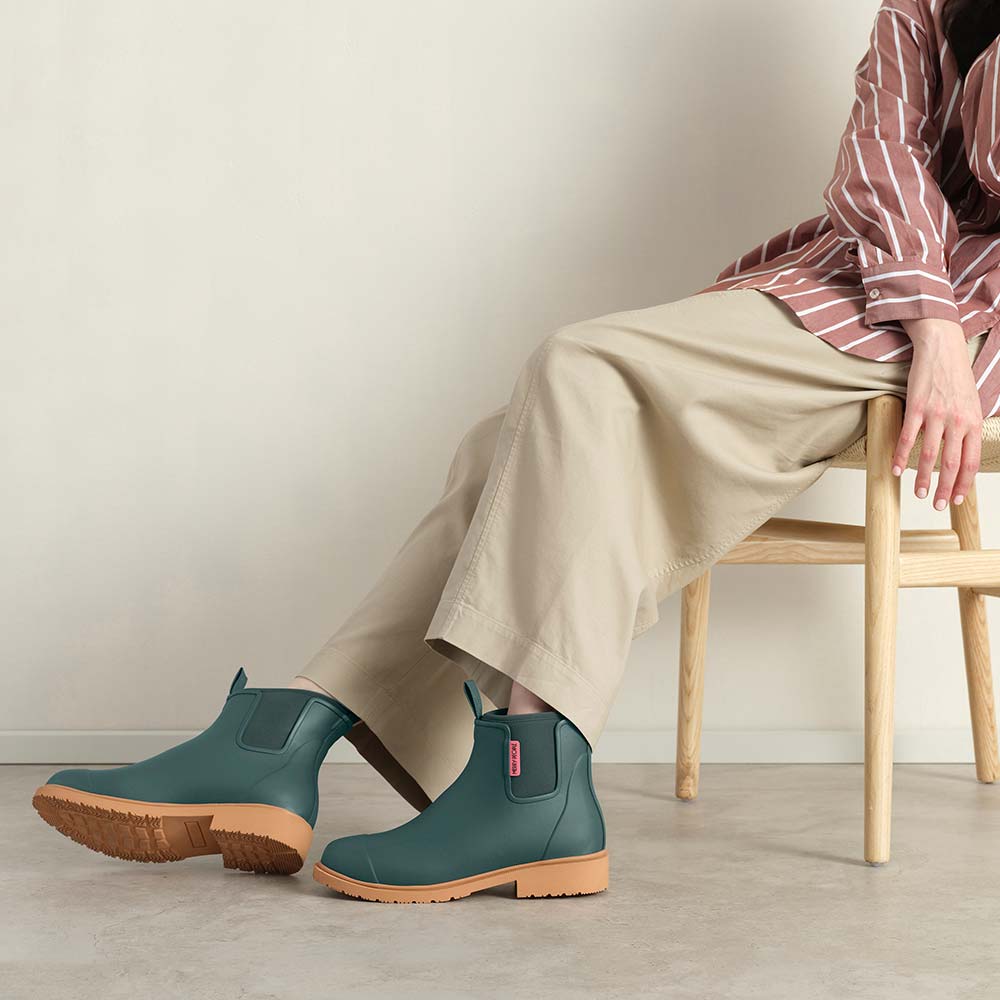 Person wearing teal rubber boots with brown soles sitting on a wooden stool.