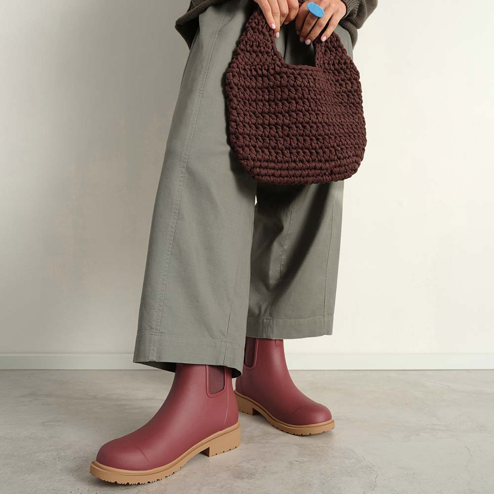 Person wearing maroon rain boots and holding a brown knitted bag against a neutral background