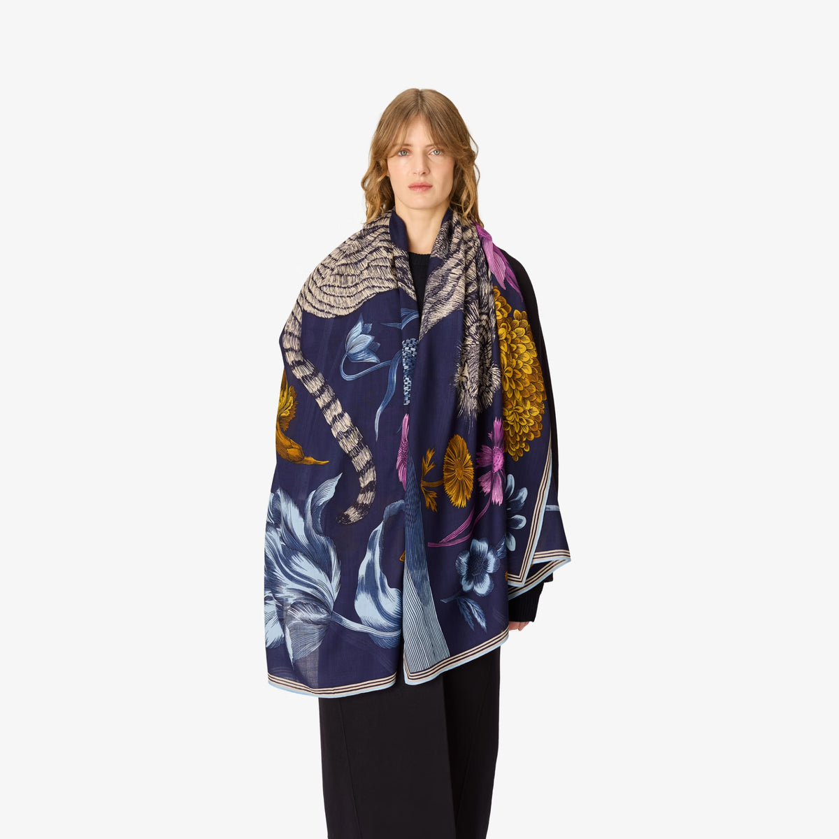 A model wearing a navy blue silk scarf with a detailed multicolor print of snakes, flowers, and leaves, styled over a black outfit against a light gray background.