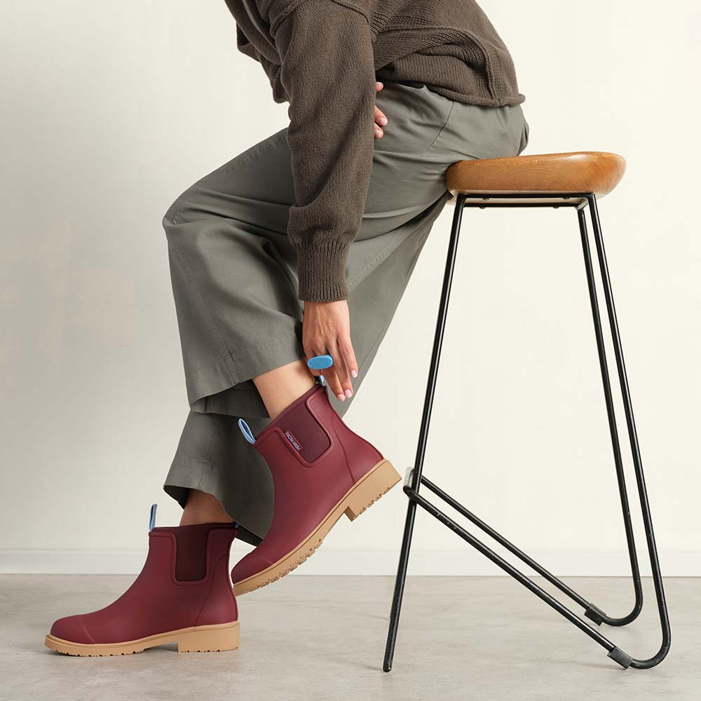Person wearing maroon boots with beige soles, sitting on a black metal stool against a plain background.