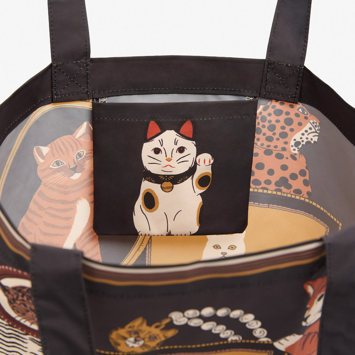 Close-up of a tote bag interior with a black front pocket featuring a white maneki-neko cat illustration, and a printed cat-pattern lining in orange, beige, and black.