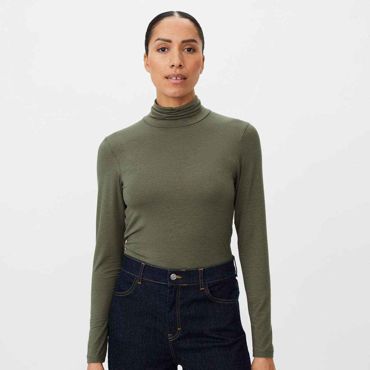 A woman modeling a fitted olive green long-sleeve turtleneck top paired with dark blue jeans, photographed against a clean light gray studio background.