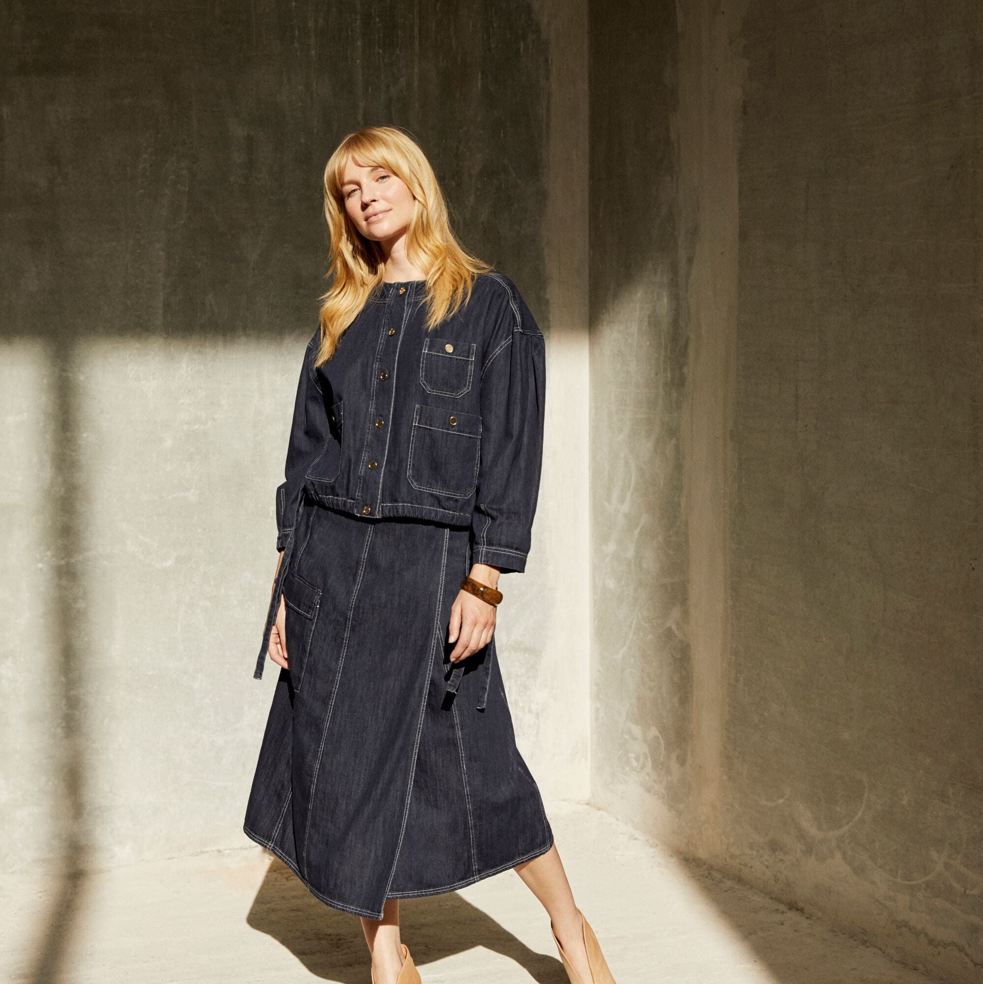 Woman wearing a dark denim outfit standing in a sunlit room with shadows on the wall.