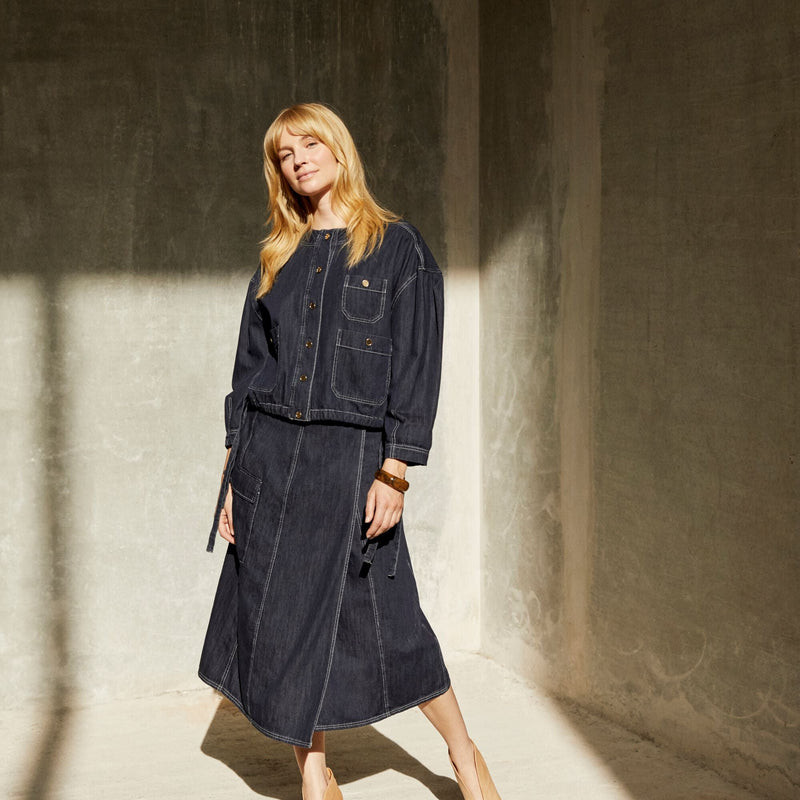 Woman wearing a dark denim outfit standing in a sunlit room with shadows on the wall.