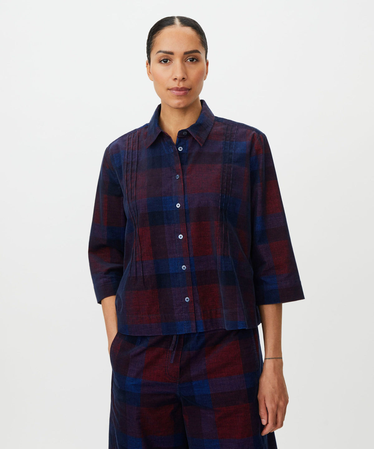 A model wearing a matching plaid button-up shirt and trousers set in dark navy and burgundy. The shirt features a relaxed fit, three-quarter sleeves, and vertical pleats on the front.