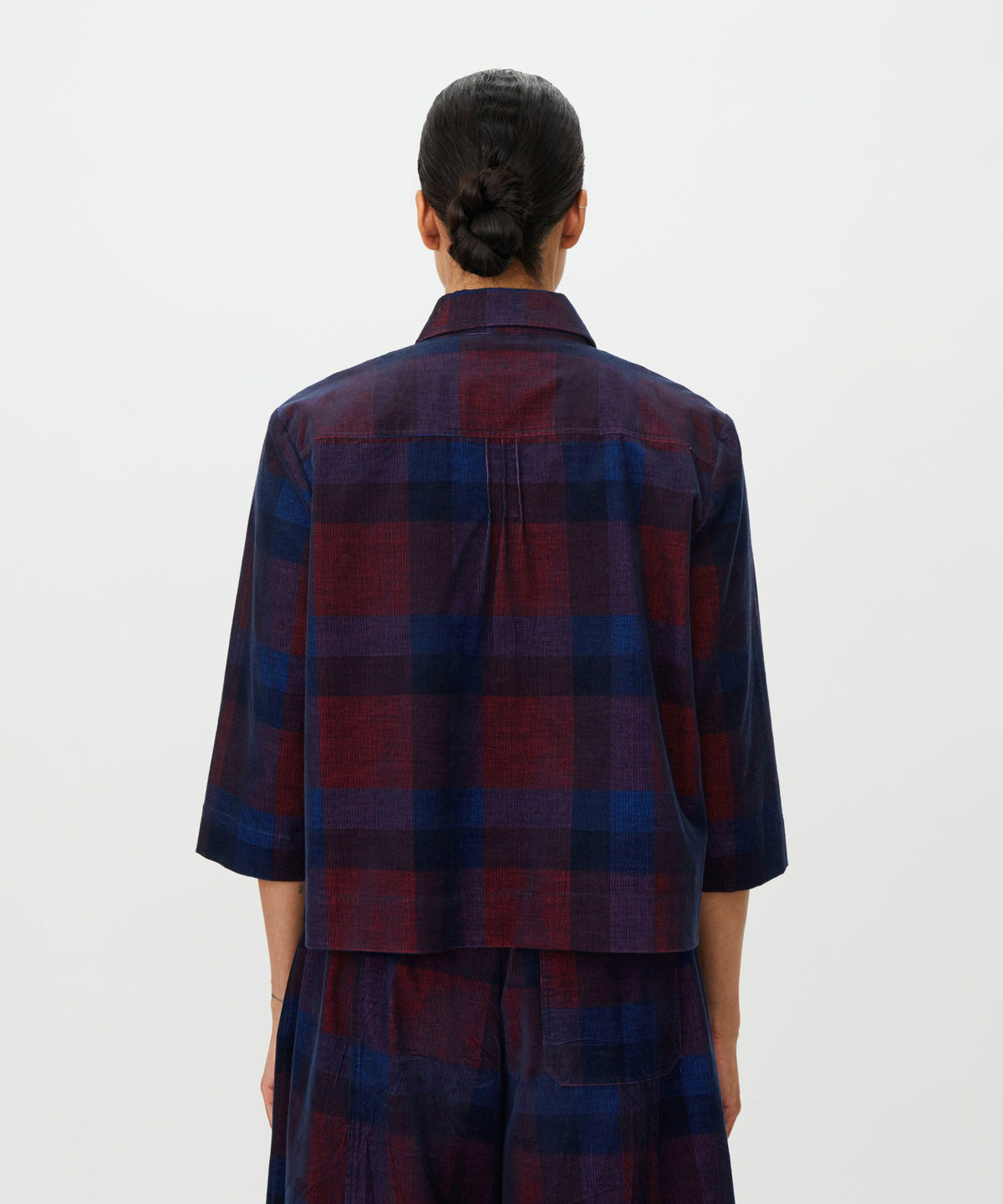 A person viewed from the back wearing a loose-fitting plaid shirt and matching pants in dark navy and burgundy red, against a plain light gray studio background.