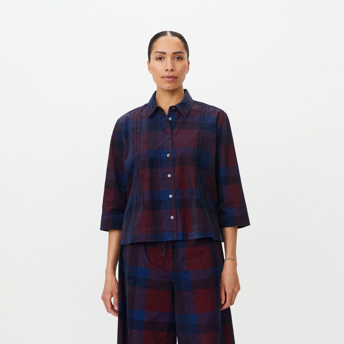 A model stands against a plain white studio background wearing a matching plaid shirt and wide-leg pants set in dark blue and burgundy, paired with black loafers.