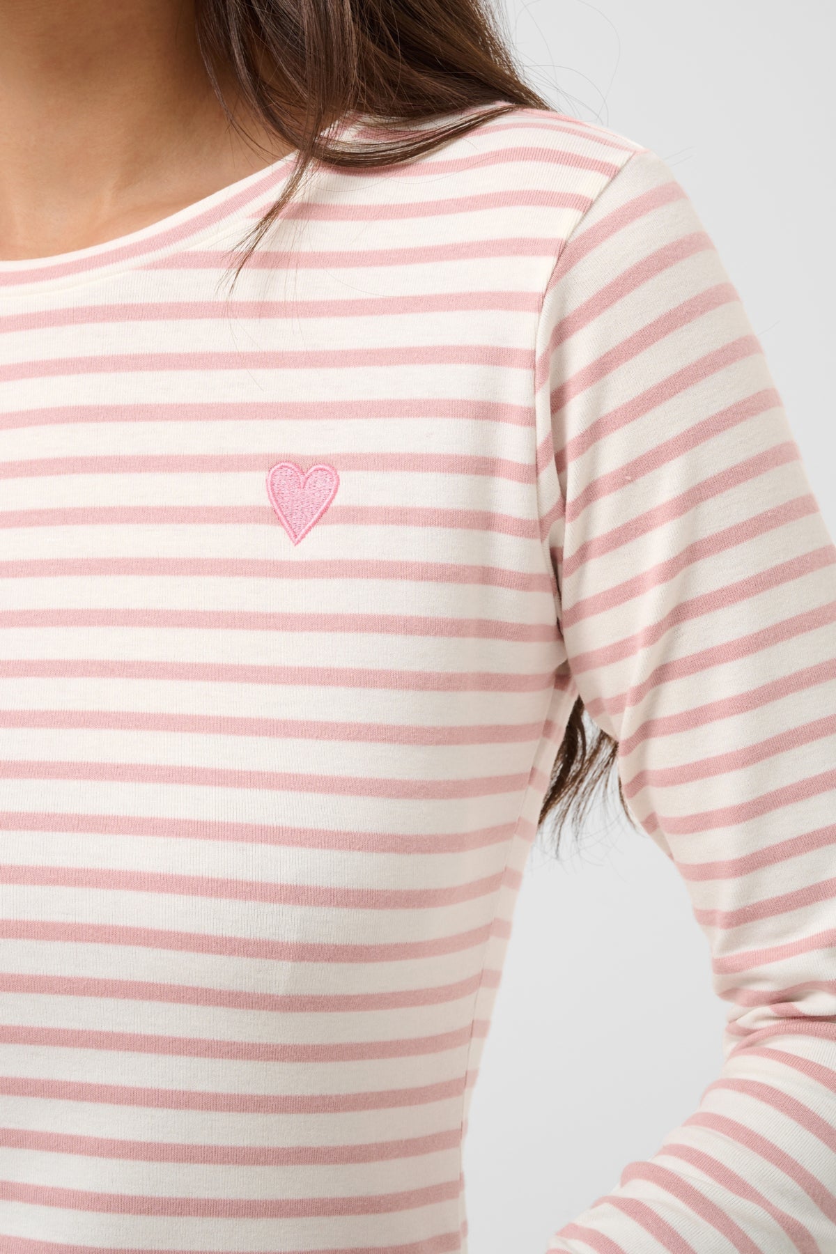A close-up of a person wearing a long-sleeve top with horizontal pink and off-white stripes and a small pink heart embroidered on the left chest, against a light gray background.