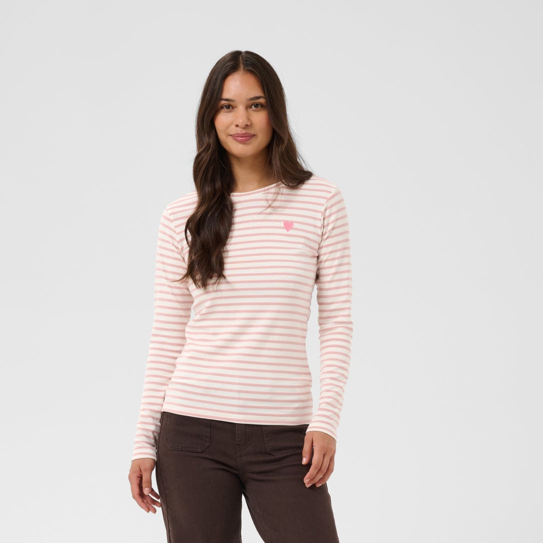 A woman modeling a light pink and white striped long-sleeve top with a small pink embroidered logo on the left chest, paired with dark brown pants and beige shoes, against a light gray studio background.