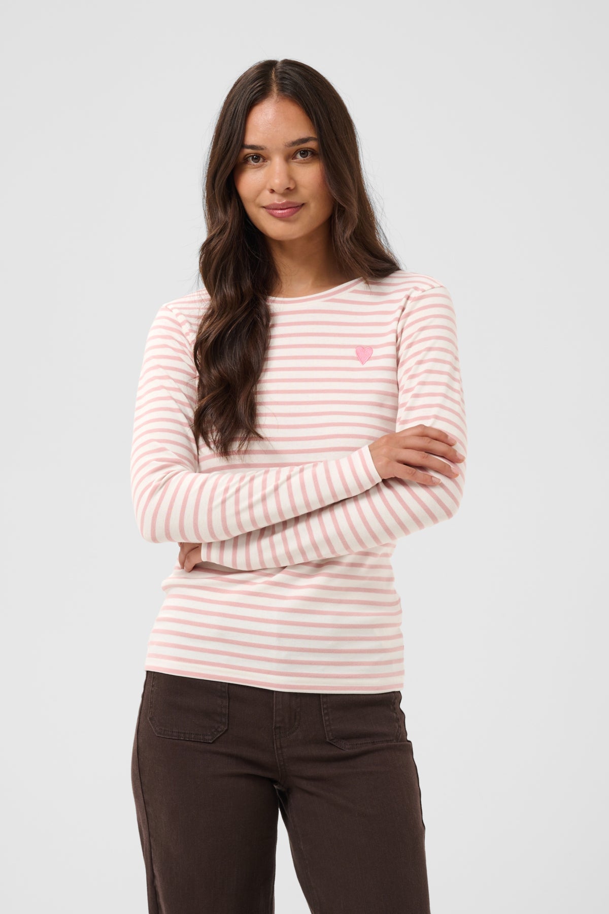 A woman modeling a light pink and white striped long-sleeve top with a small pink heart on the left chest, paired with dark brown pants, against a light gray background.