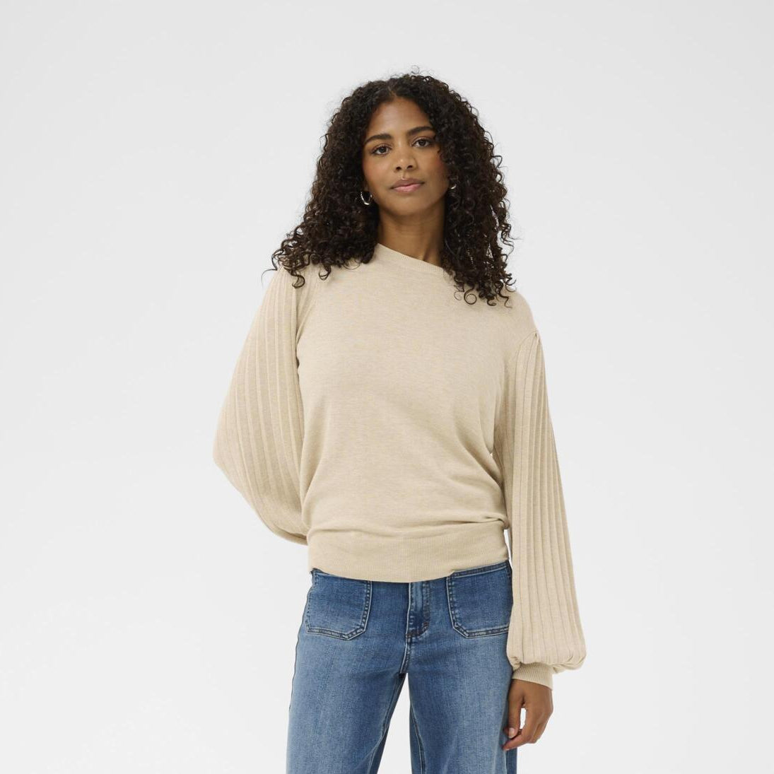 A woman with curly dark hair wearing a beige long-sleeve knit sweater with balloon sleeves and blue jeans, standing against a plain light gray studio background.