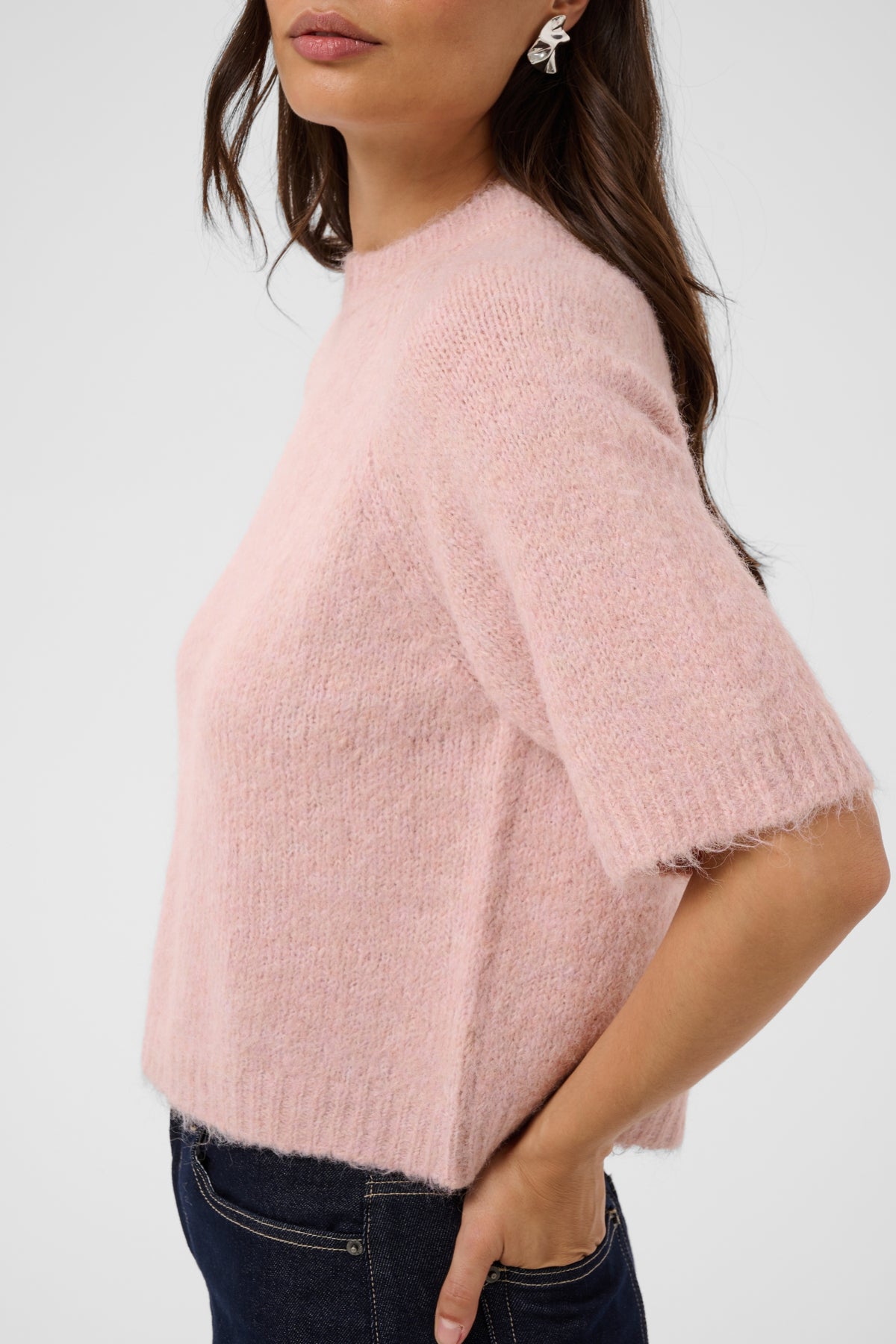 A woman wearing a fuzzy short-sleeve knit sweater in light pink, paired with dark blue jeans, shown from the side against a plain light gray background.