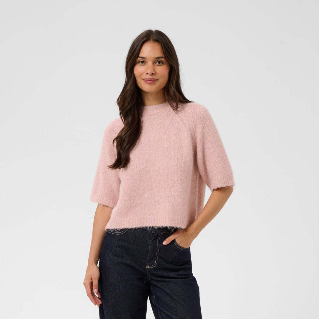 A woman modeling a light pink short-sleeve knit top and dark navy wide-leg jeans against a plain light gray studio background.