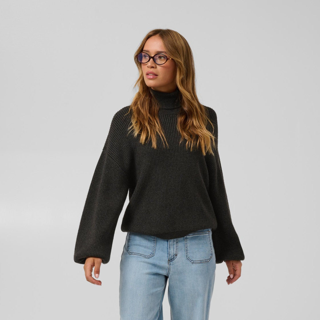 A woman modeling a black knit mock-neck sweater with wide bell sleeves, paired with light-wash blue jeans and brown pointed-toe heels, against a light gray studio background.