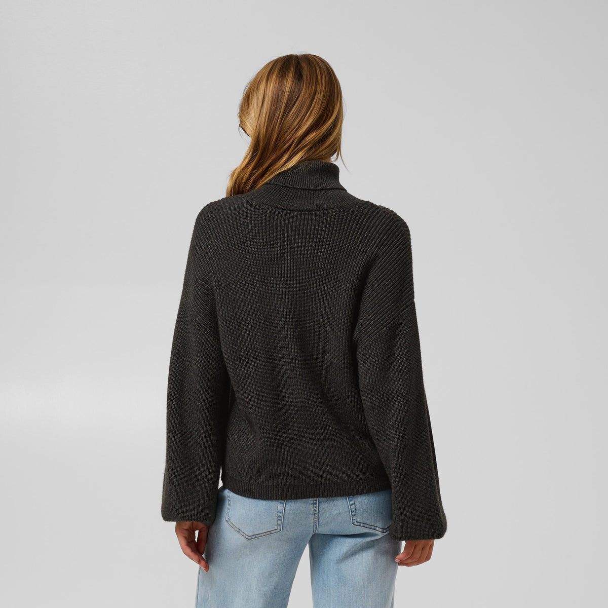 A woman is shown from the back wearing a dark charcoal gray ribbed knit sweater and light blue straight-leg jeans, standing against a light gray studio background.