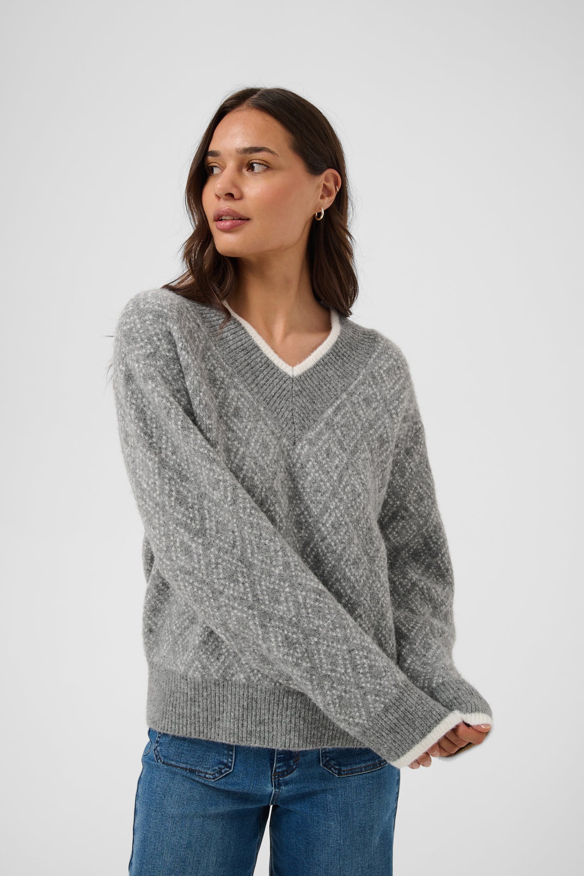 A woman wearing a gray knit sweater with a white V-neck trim and blue jeans, standing against a light gray studio background.