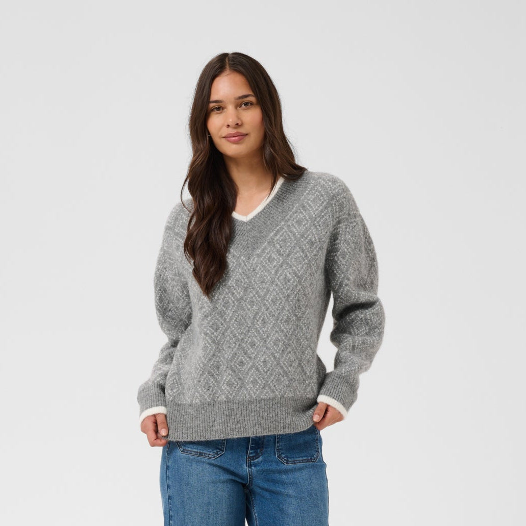 A woman modeling a gray V-neck cable-knit sweater with blue wide-leg jeans and dark brown shoes, standing against a light gray studio background.