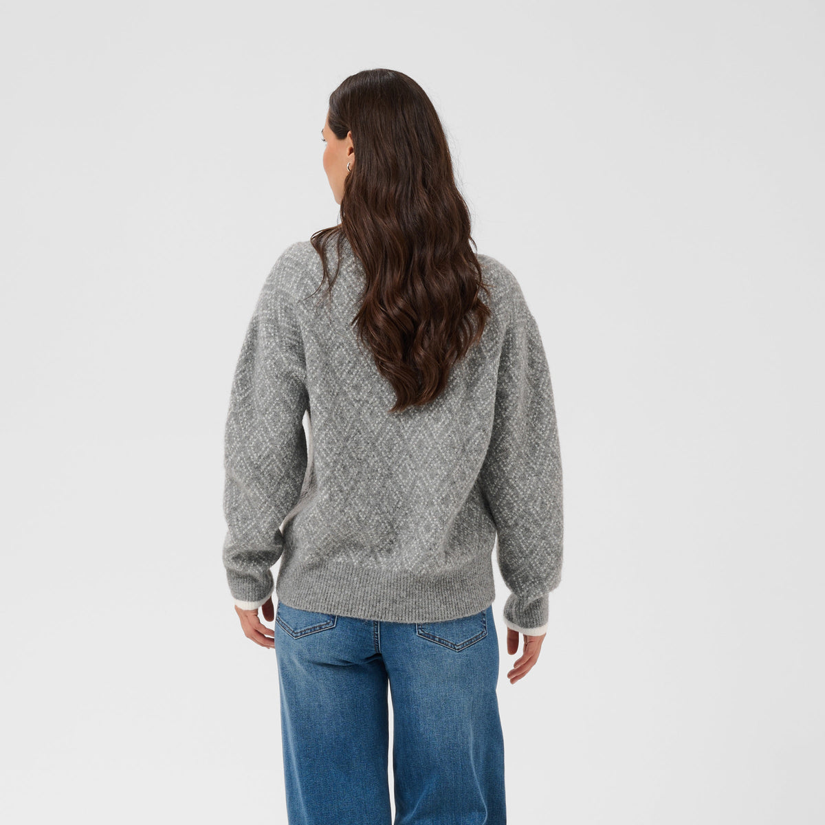A woman viewed from behind wearing a gray textured knit sweater and wide-leg blue jeans against a plain light gray studio background.