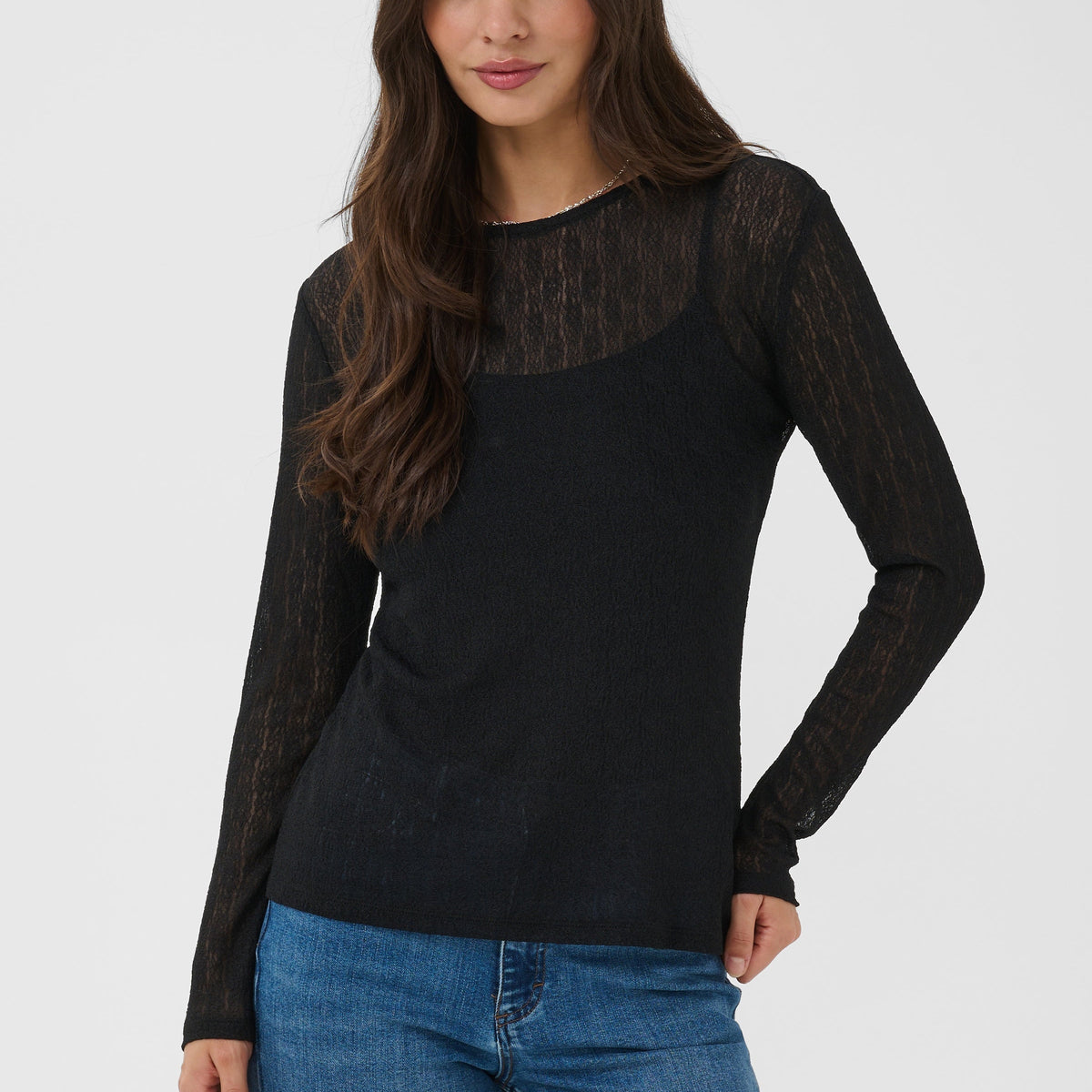 A woman modeling a black long-sleeve knit top with sheer mesh sleeves and a sheer mesh yoke, paired with blue jeans, against a light gray studio background.