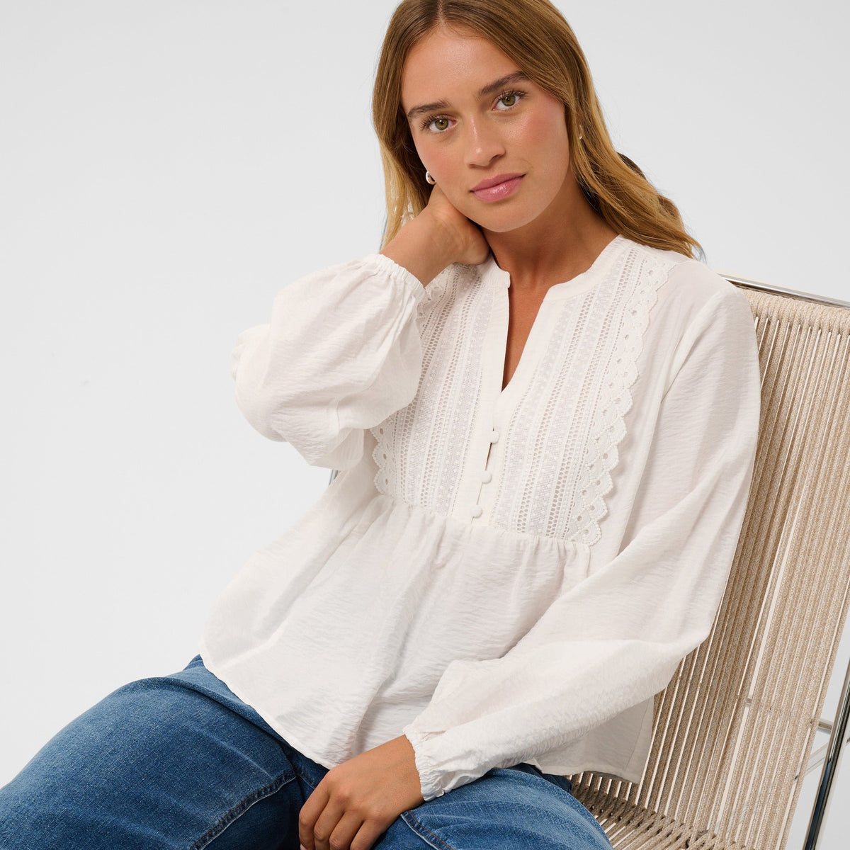 A woman wearing a white long-sleeve blouse with lace detailing and blue jeans, sitting on a woven chair against a light gray background.