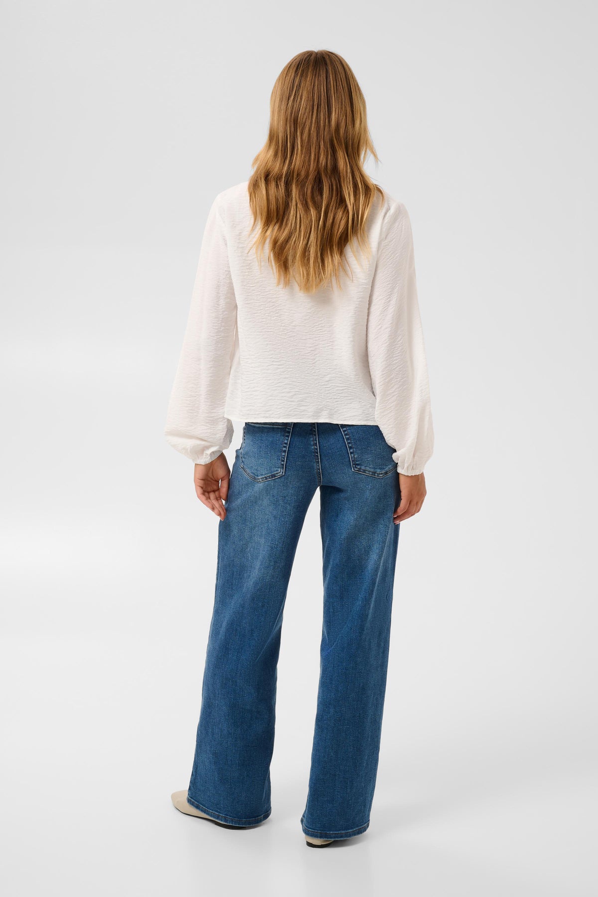 A woman viewed from behind wearing a white textured long-sleeve top and blue wide-leg jeans, standing against a light gray studio background.