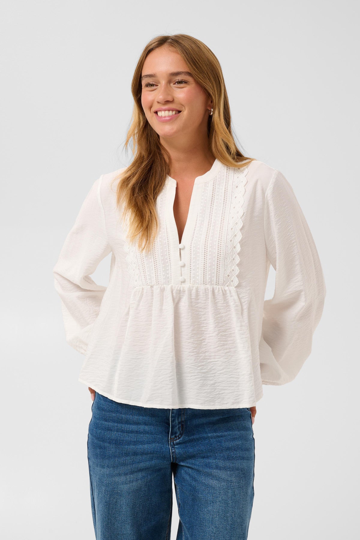 A woman wearing a white long-sleeve blouse with a split neckline, decorative scalloped embroidery, and a gathered waist, paired with blue jeans, against a light gray studio background.