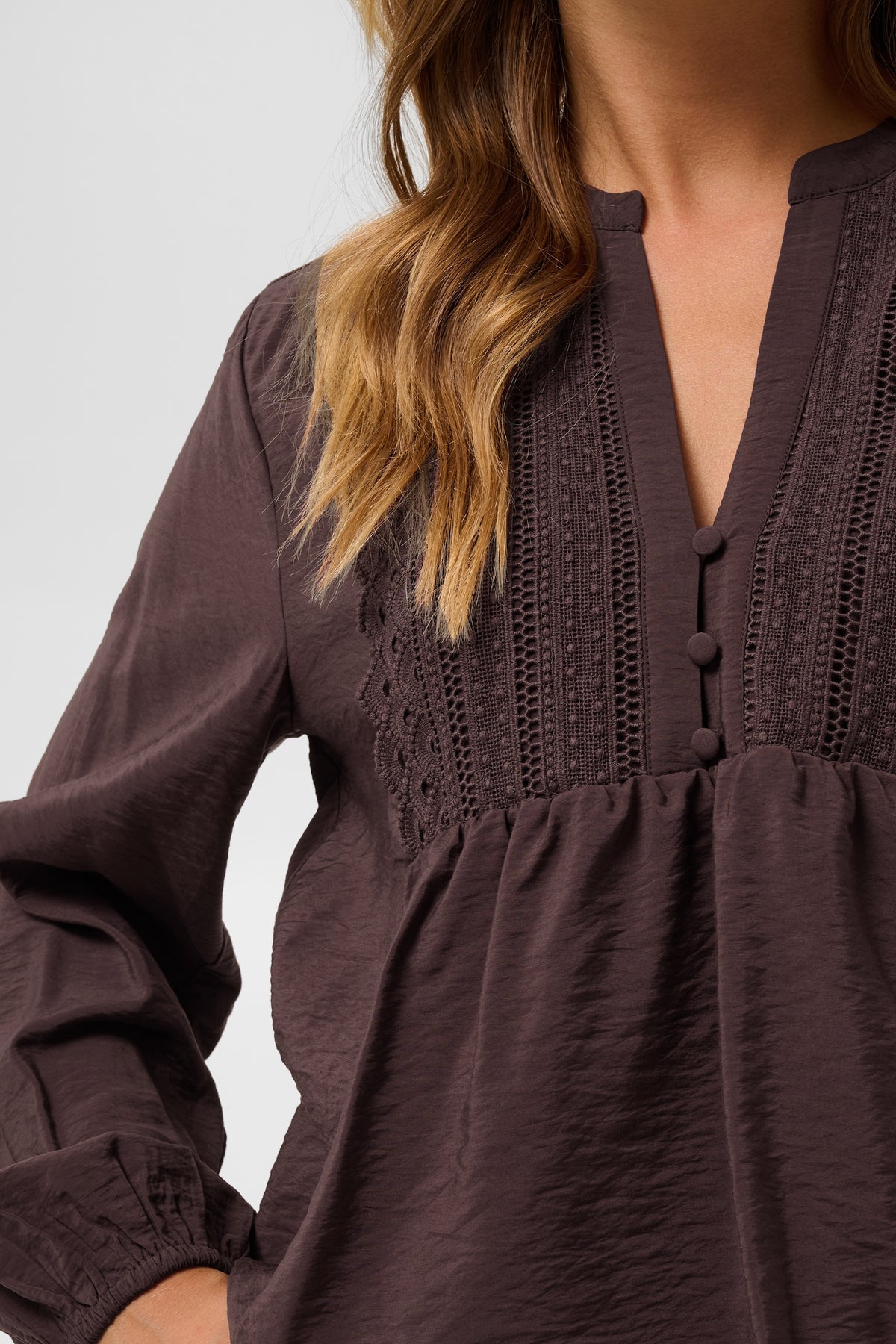 A close-up of a woman wearing a dark brown long-sleeve blouse with a split V-neckline, decorative eyelet embroidery, and a gathered waist seam.