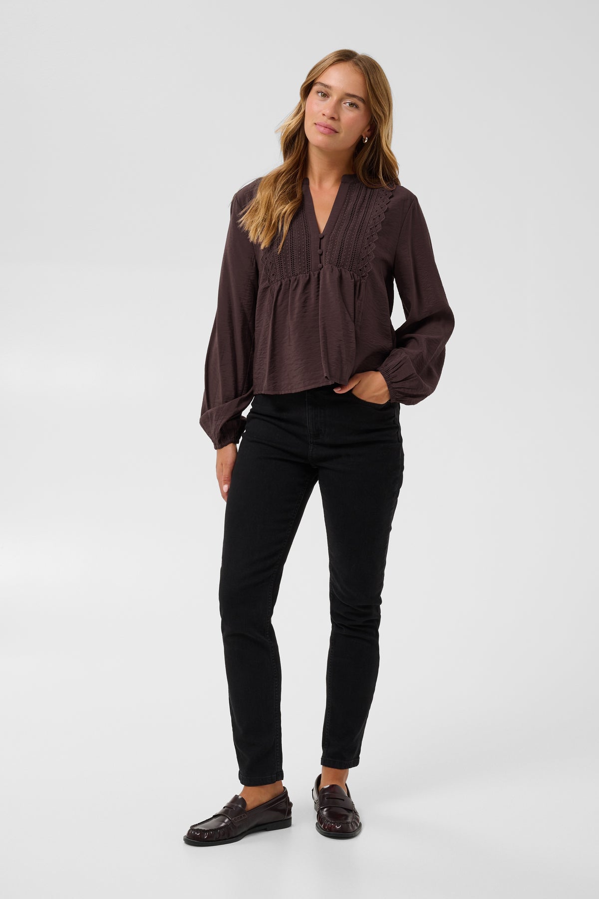 A woman modeling a dark brown long-sleeve blouse with a gathered peplum waist and ruffled trim, paired with black pants and brown loafers, against a light gray studio background.