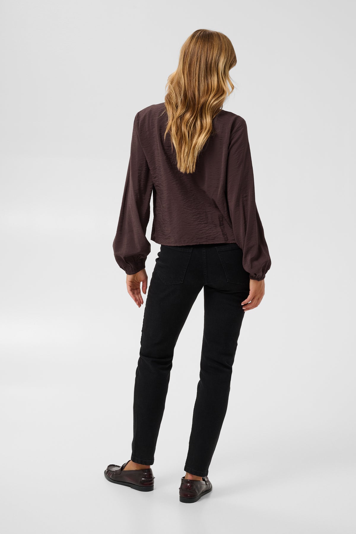A woman is shown from behind wearing a dark brown long-sleeve blouse and black pants against a light gray studio background.