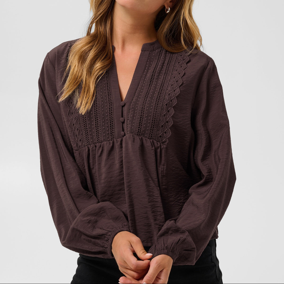 A woman modeling a dark brown long-sleeve blouse with a gathered, scalloped V-neckline and textured fabric, paired with black pants, against a light gray studio background.