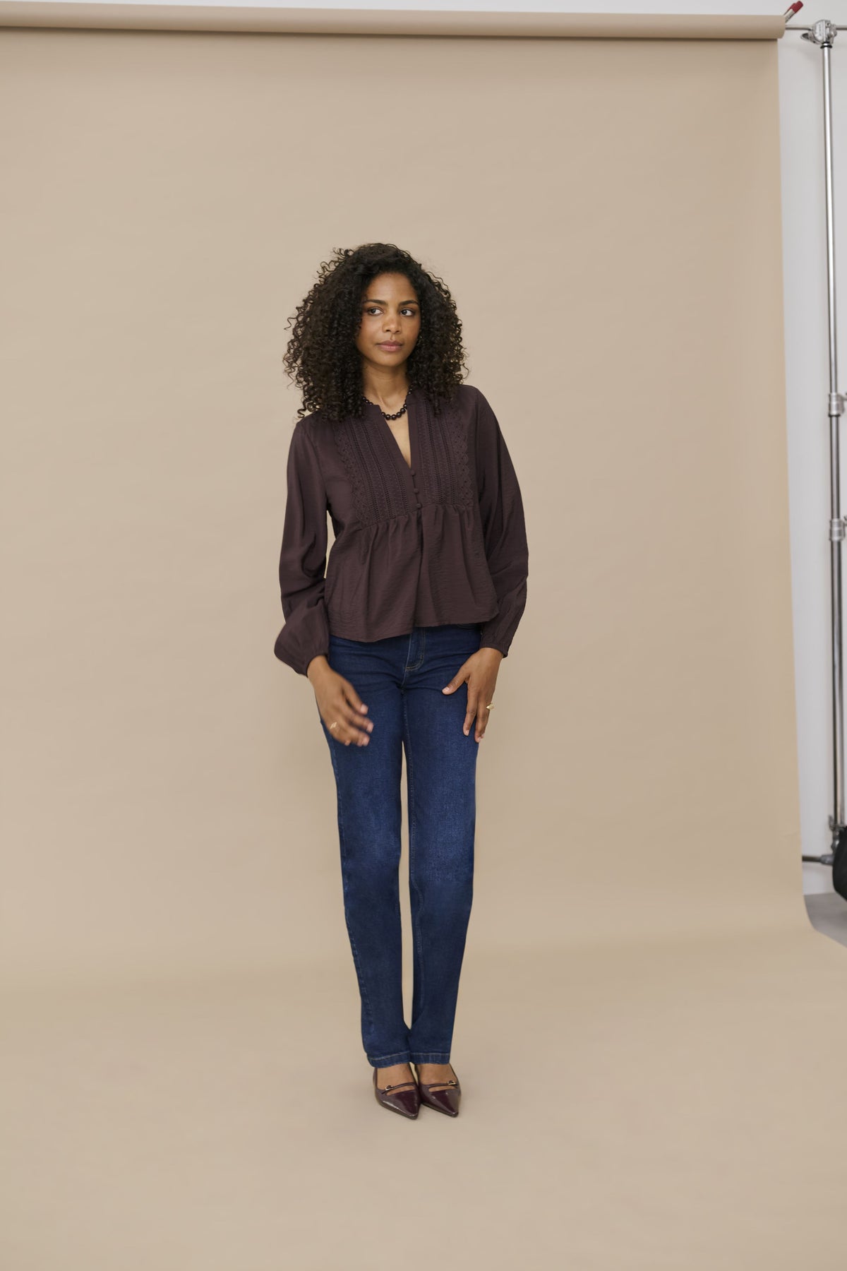 A woman with curly dark hair stands in a studio wearing a dark brown long-sleeve peplum blouse, blue jeans, and dark brown pointed flats against a beige backdrop.