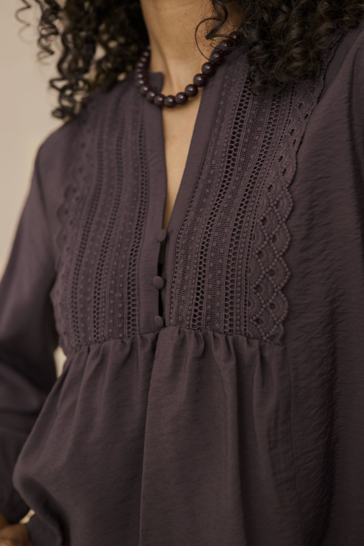 Close-up of a woman wearing a dark plum purple blouse with intricate crochet and eyelet embroidery on the chest, featuring a split neckline and gathered waist.