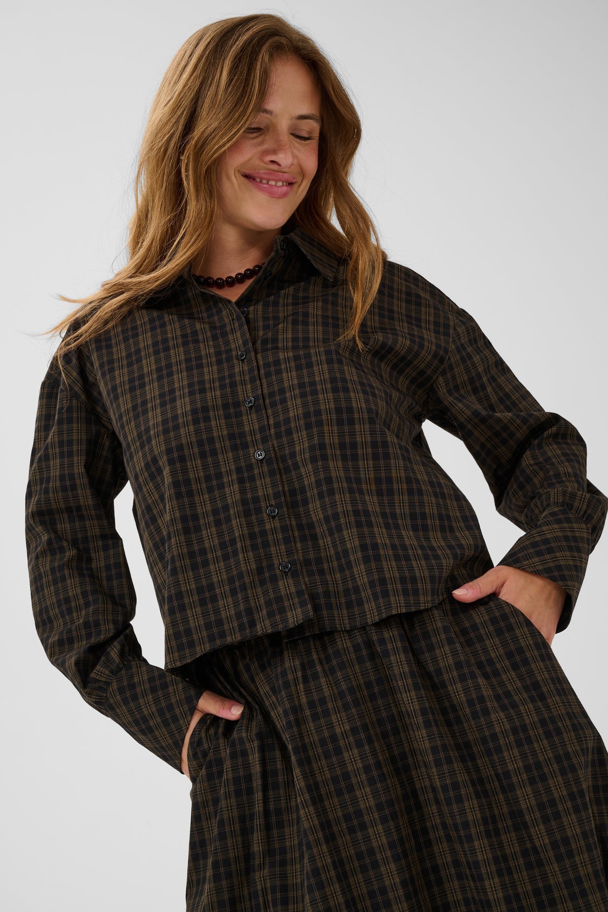 A woman wearing a dark brown and black plaid button-up shirt and matching skirt, posing with her hands in her pockets against a light gray background.